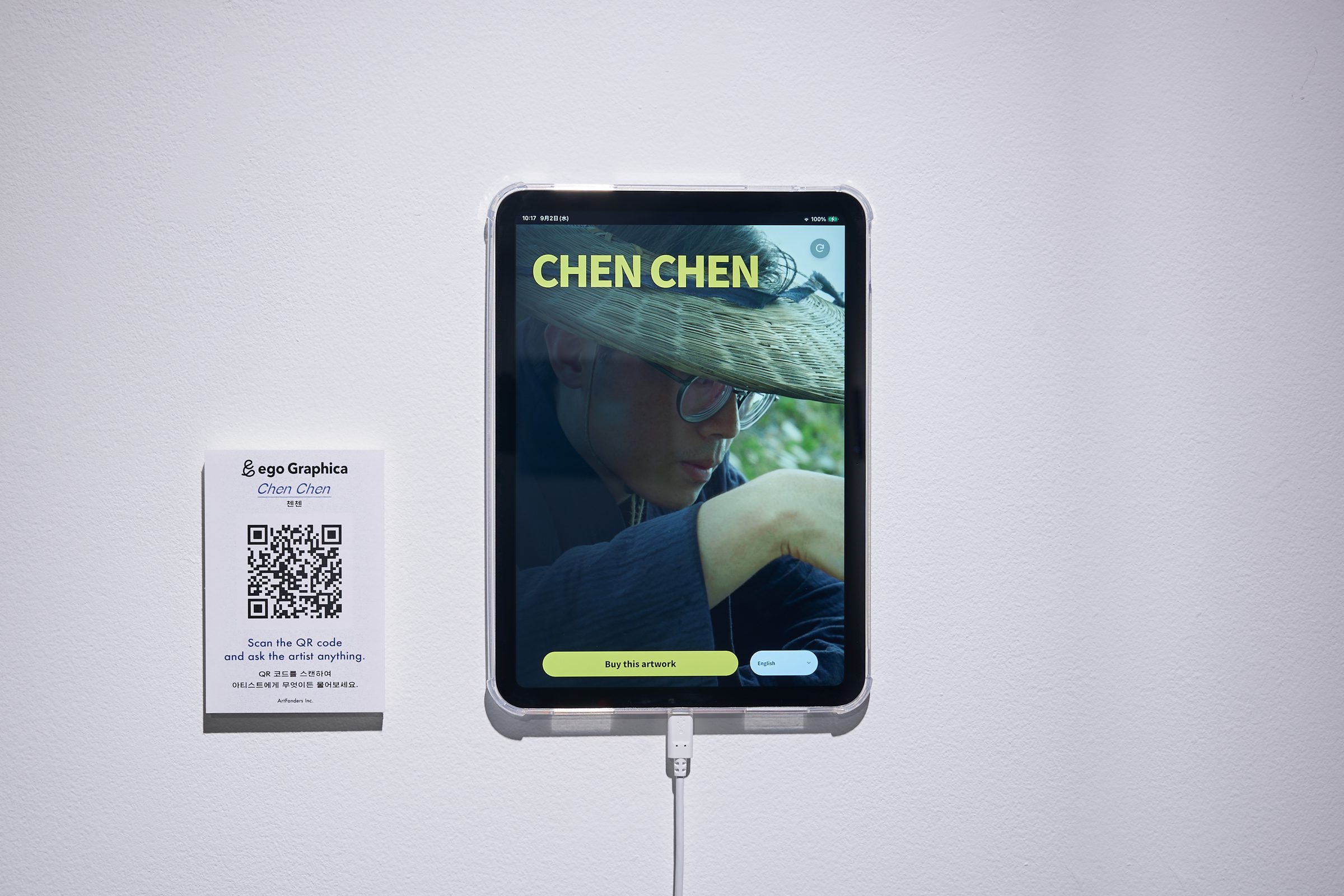 Exhibition view: an artwork on a table beside a wall-mounted iPad running the artist AI, with a QR code panel for visitors to scan.